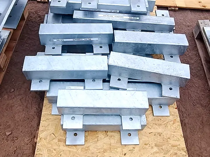 Soportes metálicos galvanizados con placas base perforadas apilados sobre palet en patio de tierra